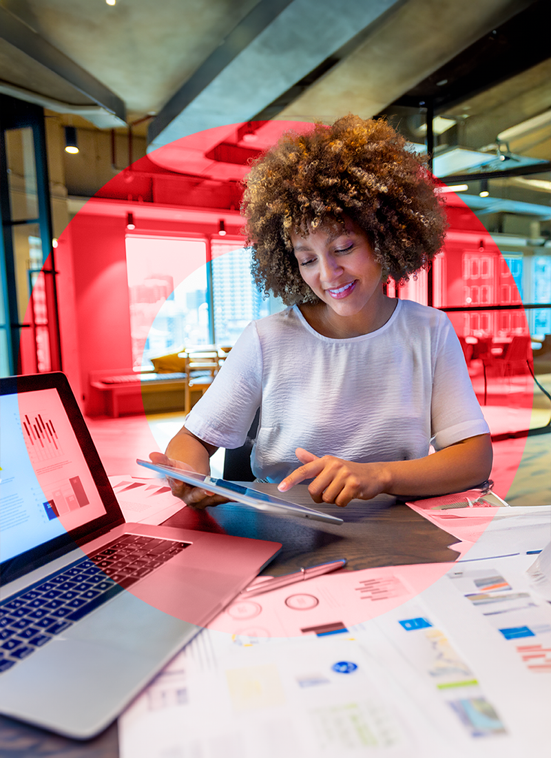 Woman in office smiling and sitting at table holding tablet next to laptop on table.